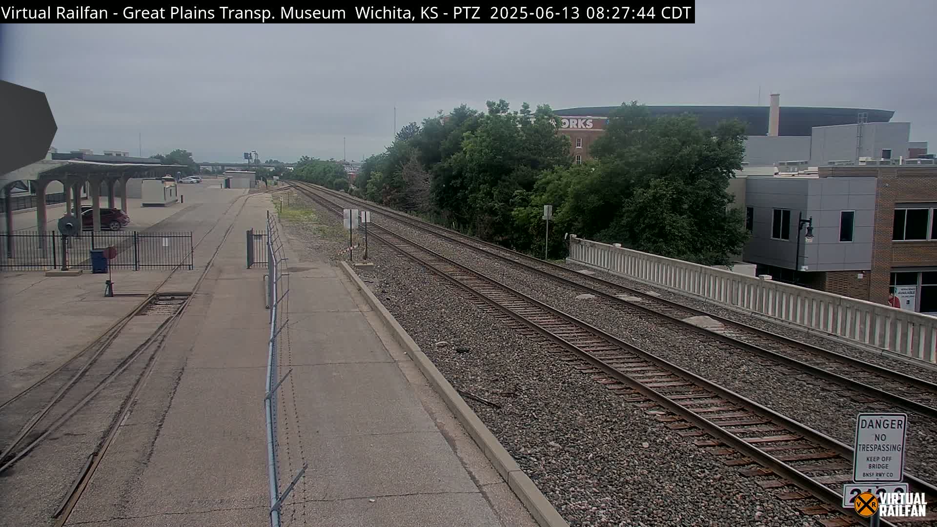 A mostly overcast day reveals a view of railroad tracks alongside a paved road and a low concrete wall, with buildings and trees in the background.