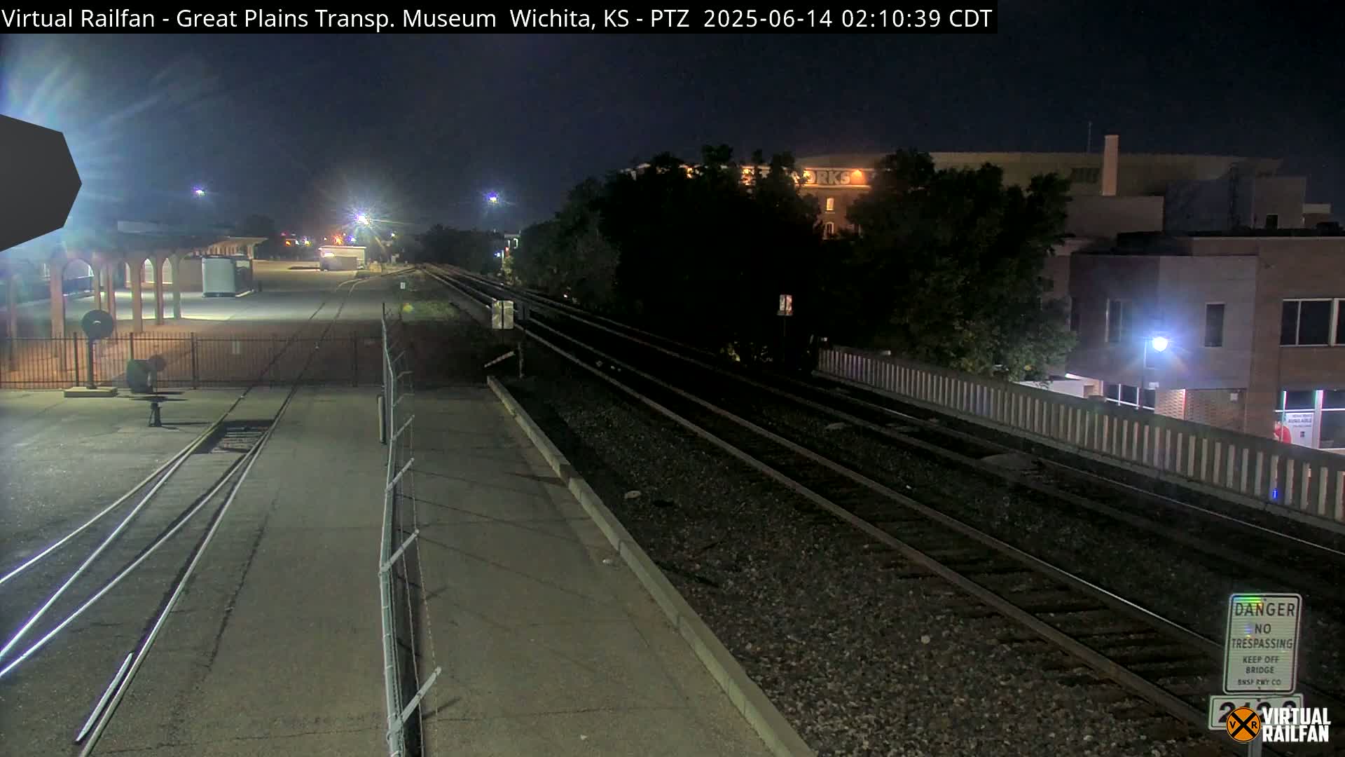 At night, a nighttime view shows train tracks extending into the distance, passing by a building and a fence, under a dark sky.