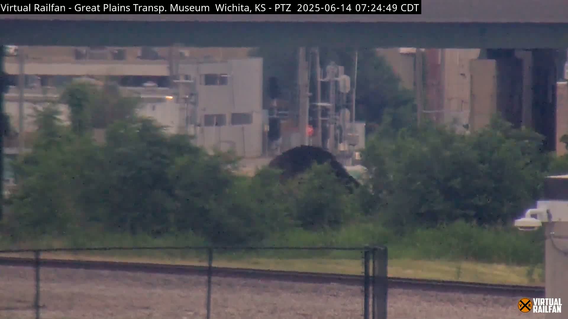 A hazy view shows a section of railroad tracks, a chain-link fence, and lush green bushes in front of industrial buildings.