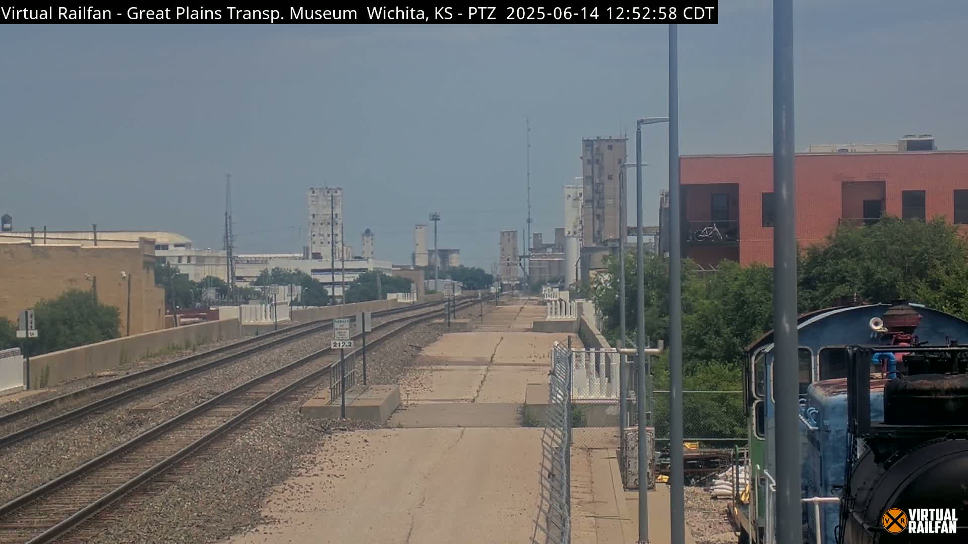 A view down a set of train tracks on a partly cloudy day, with industrial buildings and a partially visible locomotive in the foreground.