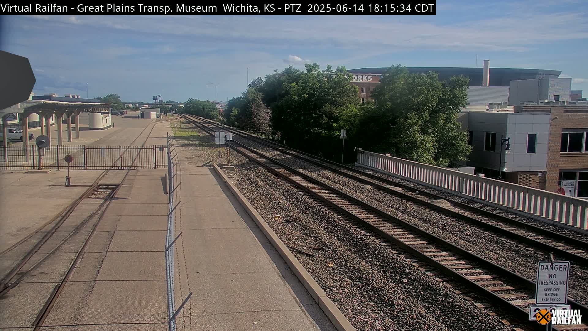 A view down a set of train tracks on a partly cloudy day, with industrial buildings and a partially visible locomotive in the foreground.
