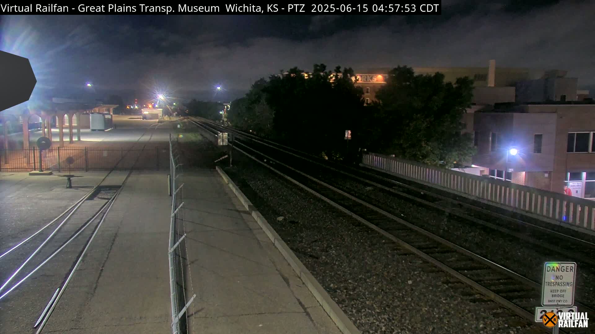 A nighttime view of train tracks next to a platform and a building, under a mostly cloudy sky.
