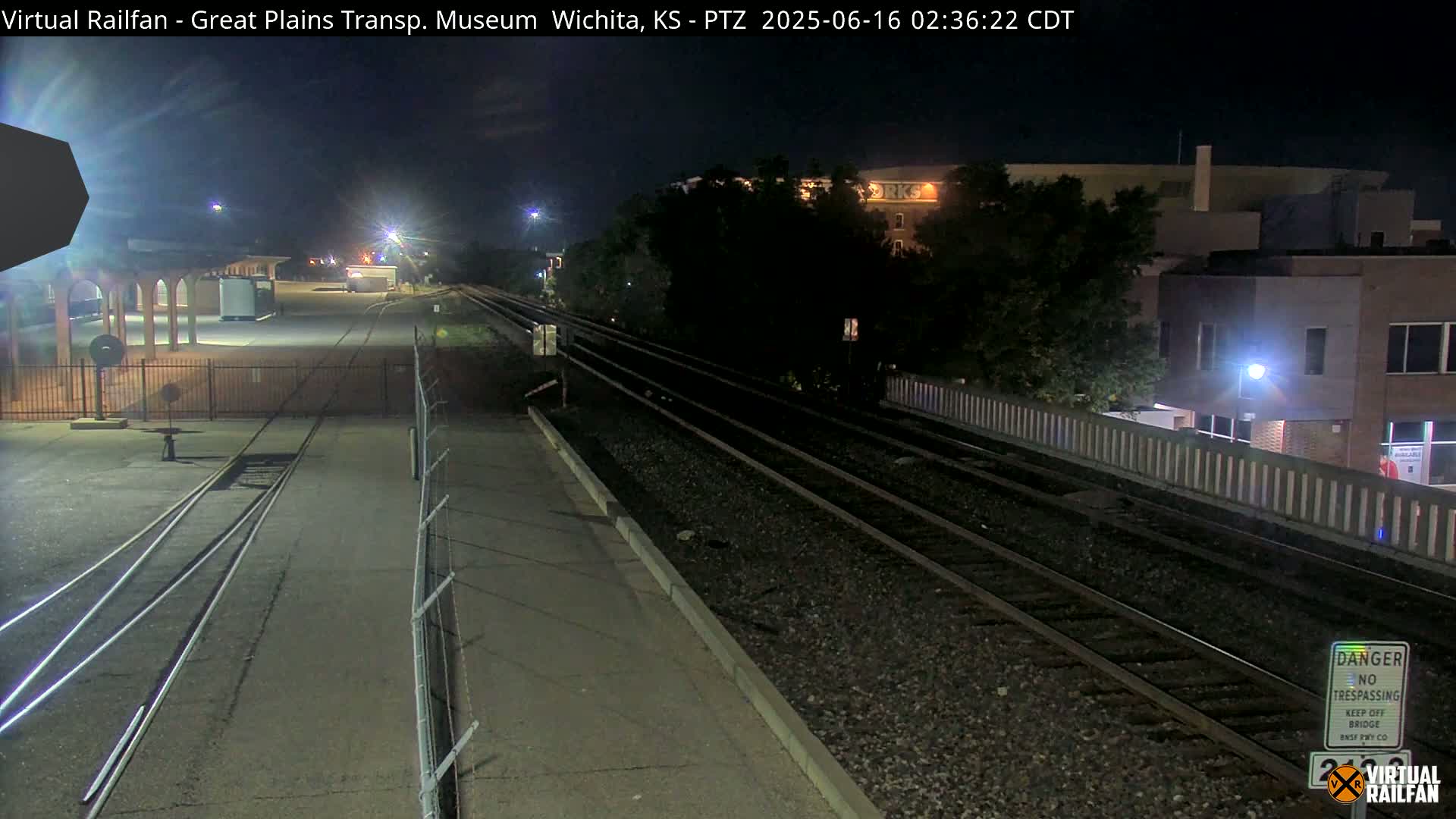 At night, a train track runs alongside a building and a paved area, under a clear sky.