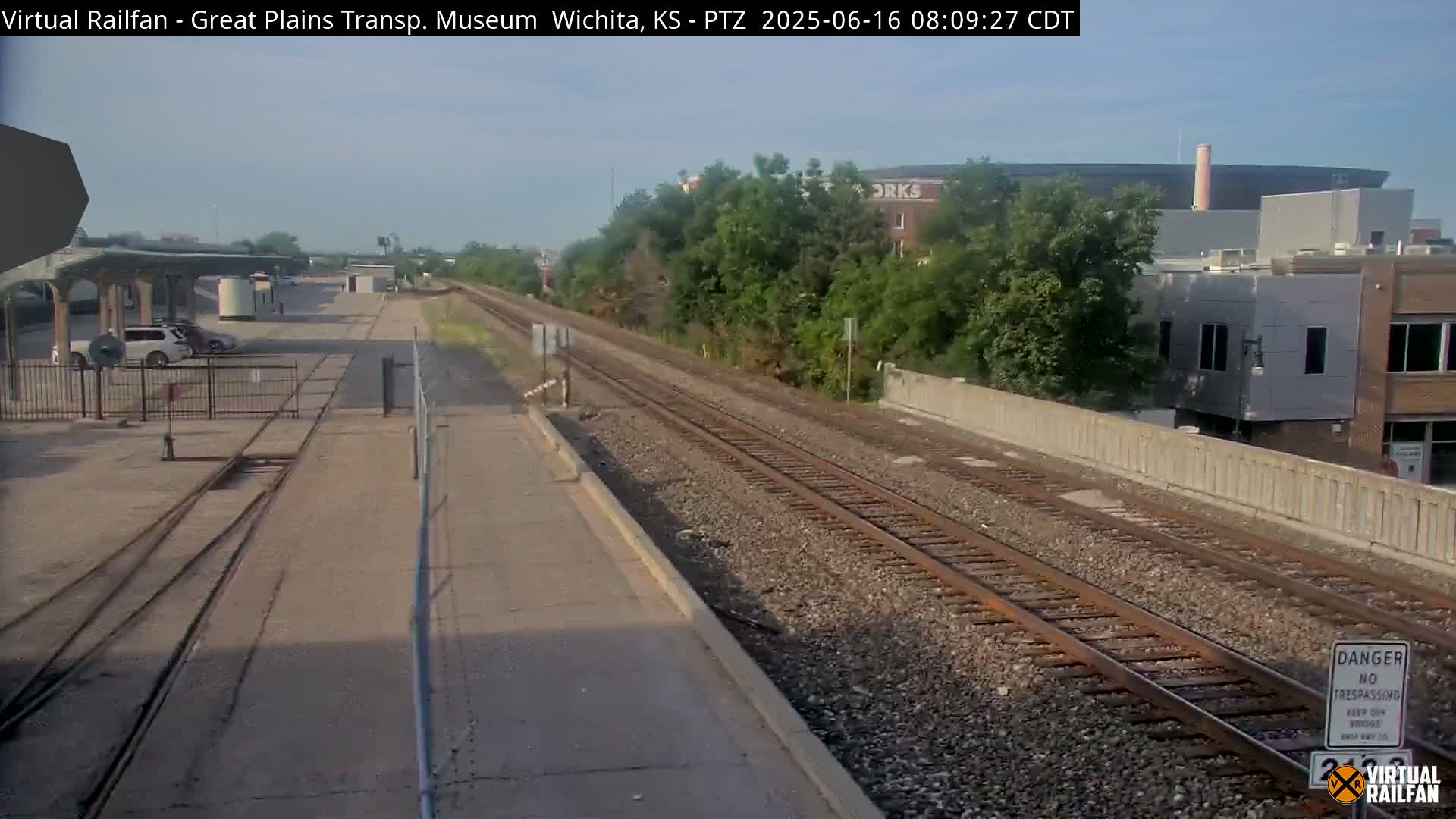A view of train tracks and a railway platform on a sunny day, with some buildings and greenery visible in the background.
