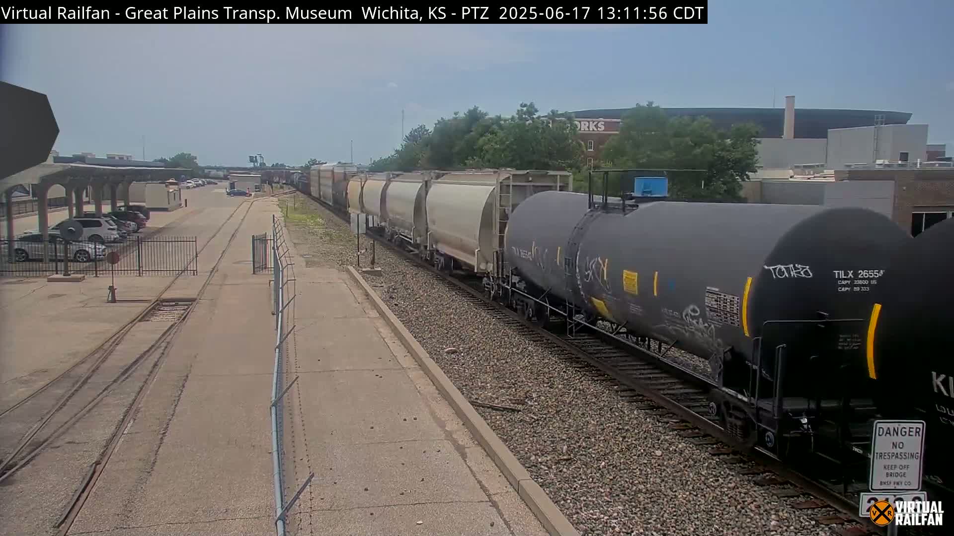 A long freight train passes by a parking lot on a sunny day.