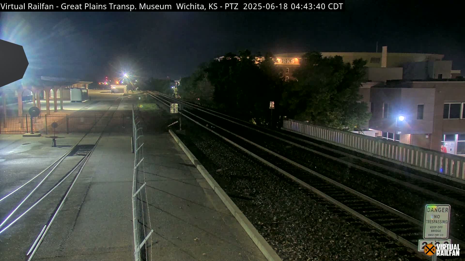 A nighttime view of railroad tracks at a station, with a building and trees visible in the background under a clear, dark sky.