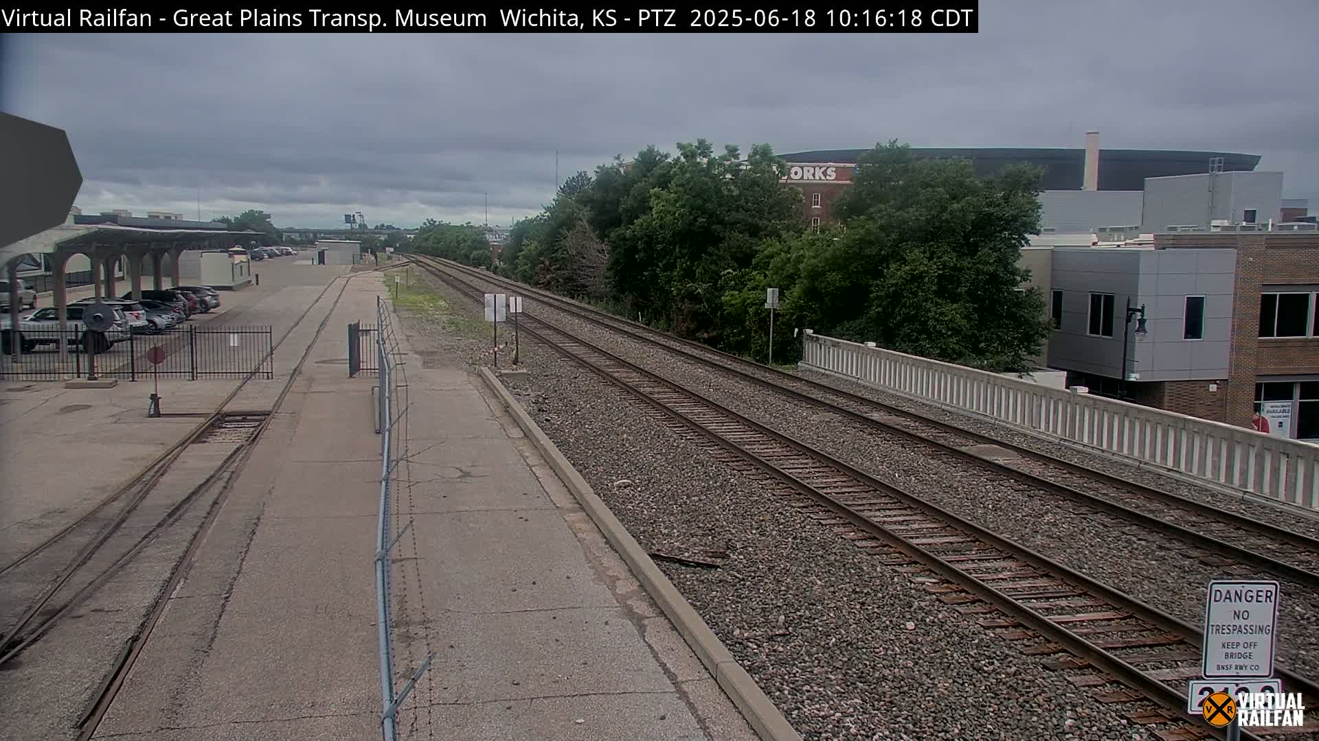 A view of railroad tracks on an overcast day, with a parking lot and buildings visible in the background.