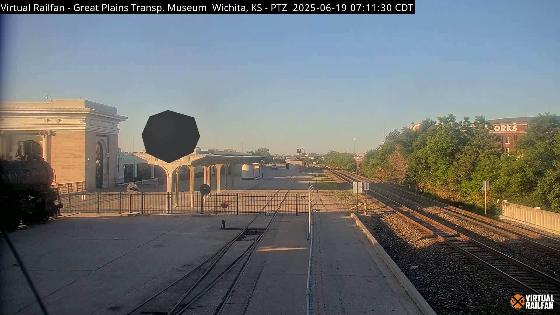 A train sits at a station platform next to railroad tracks under a clear, sunny sky.