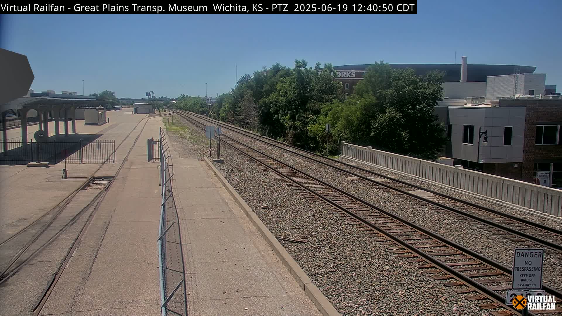 A sunny day reveals a train station platform next to railroad tracks that lead toward a tree line and buildings in the distance.