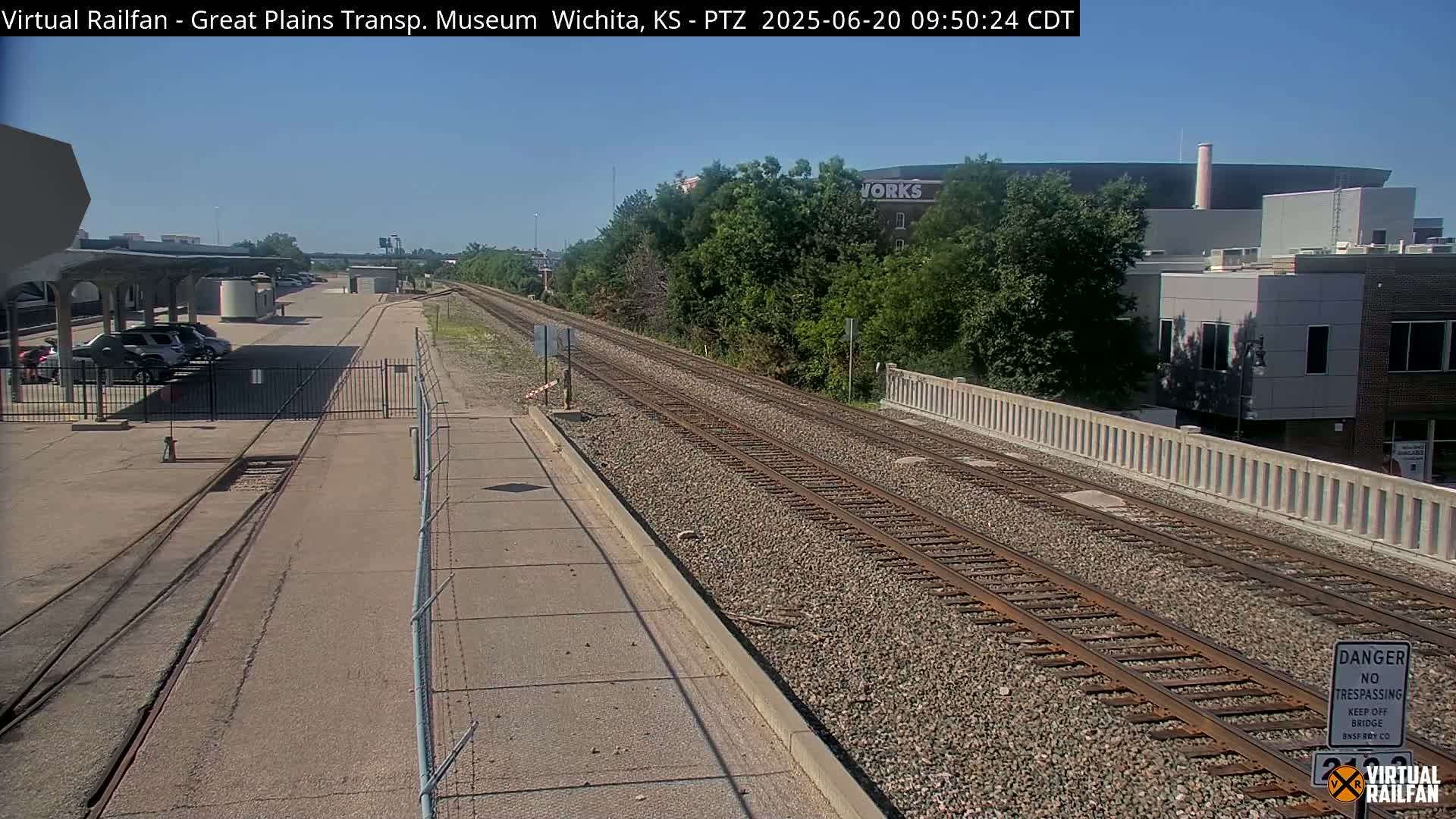 A sunny day view of railroad tracks extending from a parking lot with cars under a covered area, toward buildings and trees in the distance.