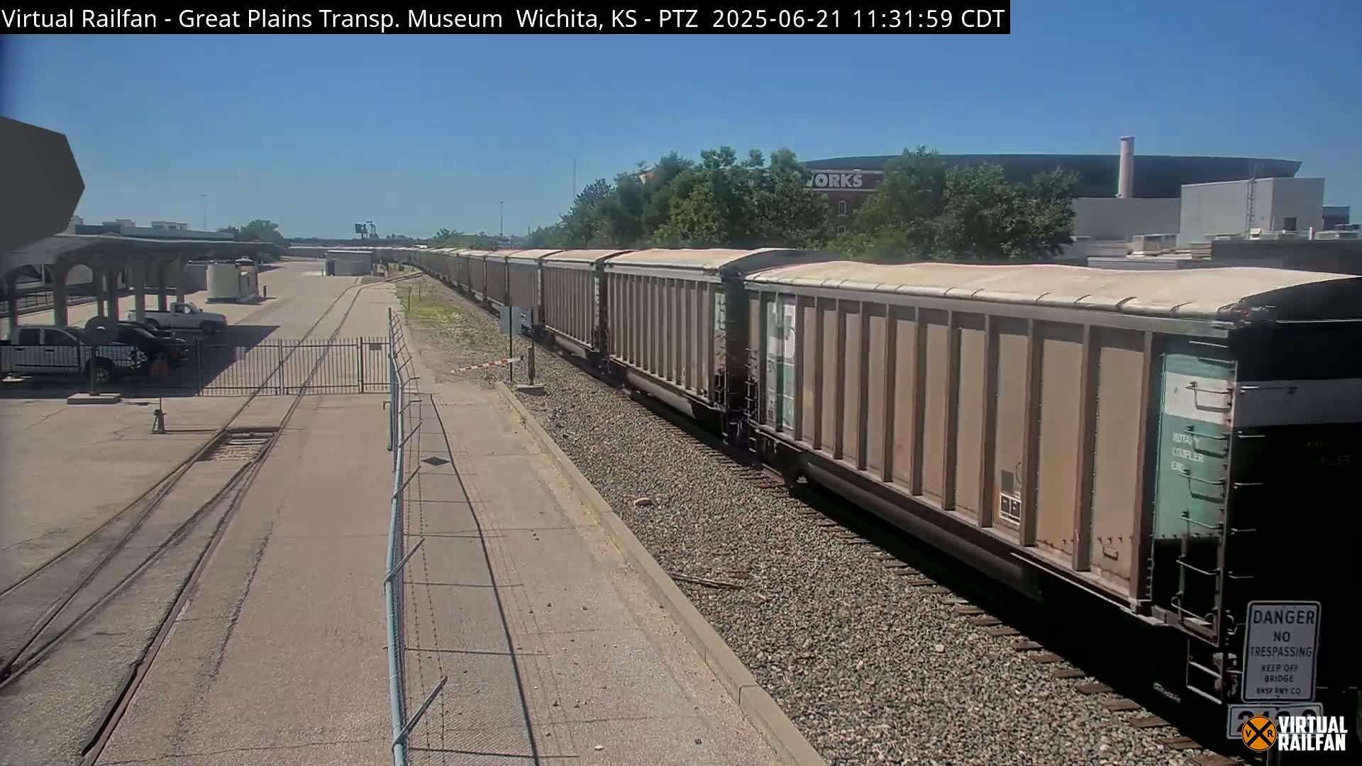 A long freight train passes by a fenced-in area under a sunny sky.