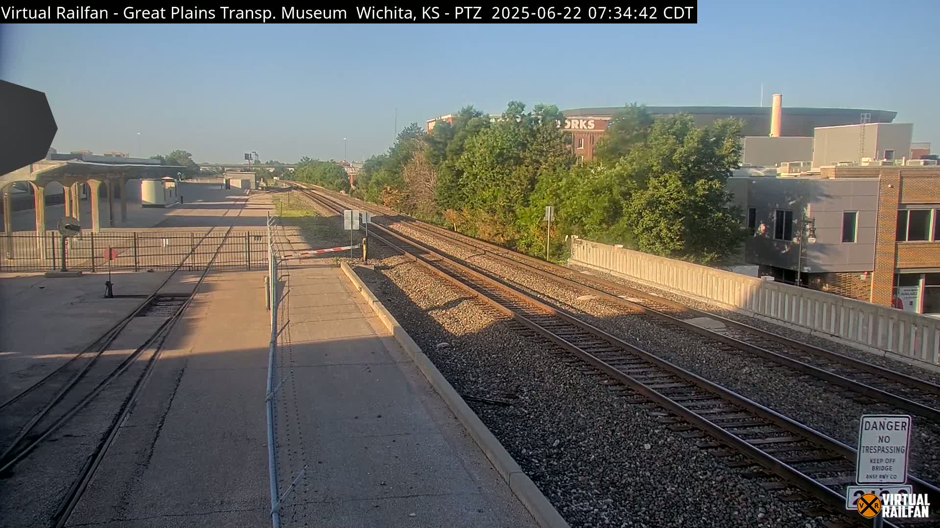 Train tracks extend into the distance under a clear sky past a fenced-in station and several buildings.