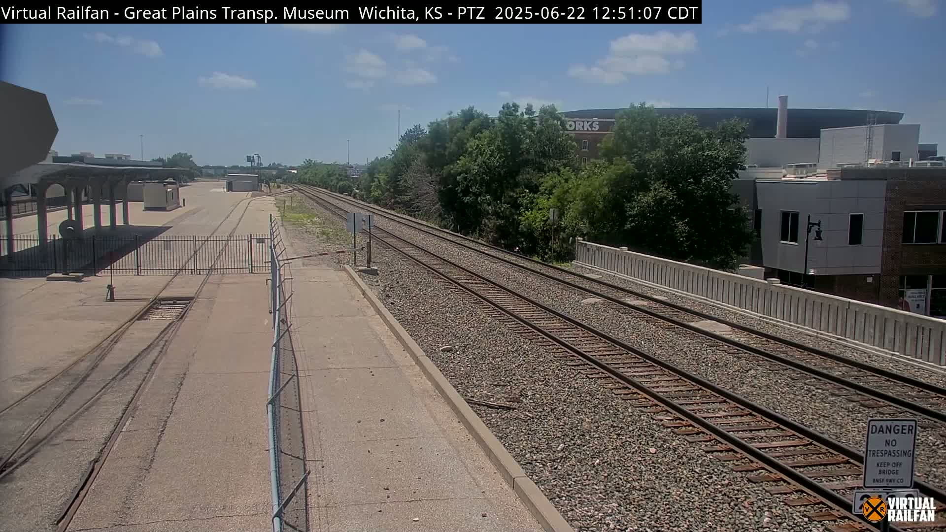 A mostly sunny day shows a view of railroad tracks extending into the distance, alongside a paved area, a chain-link fence, and buildings with trees in the background.