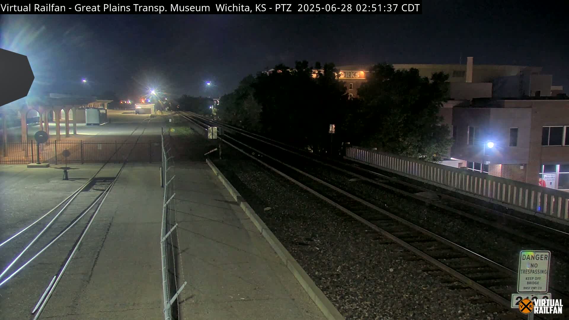 A nighttime view of railroad tracks, a fenced-in area, and a building in the distance under clear skies.