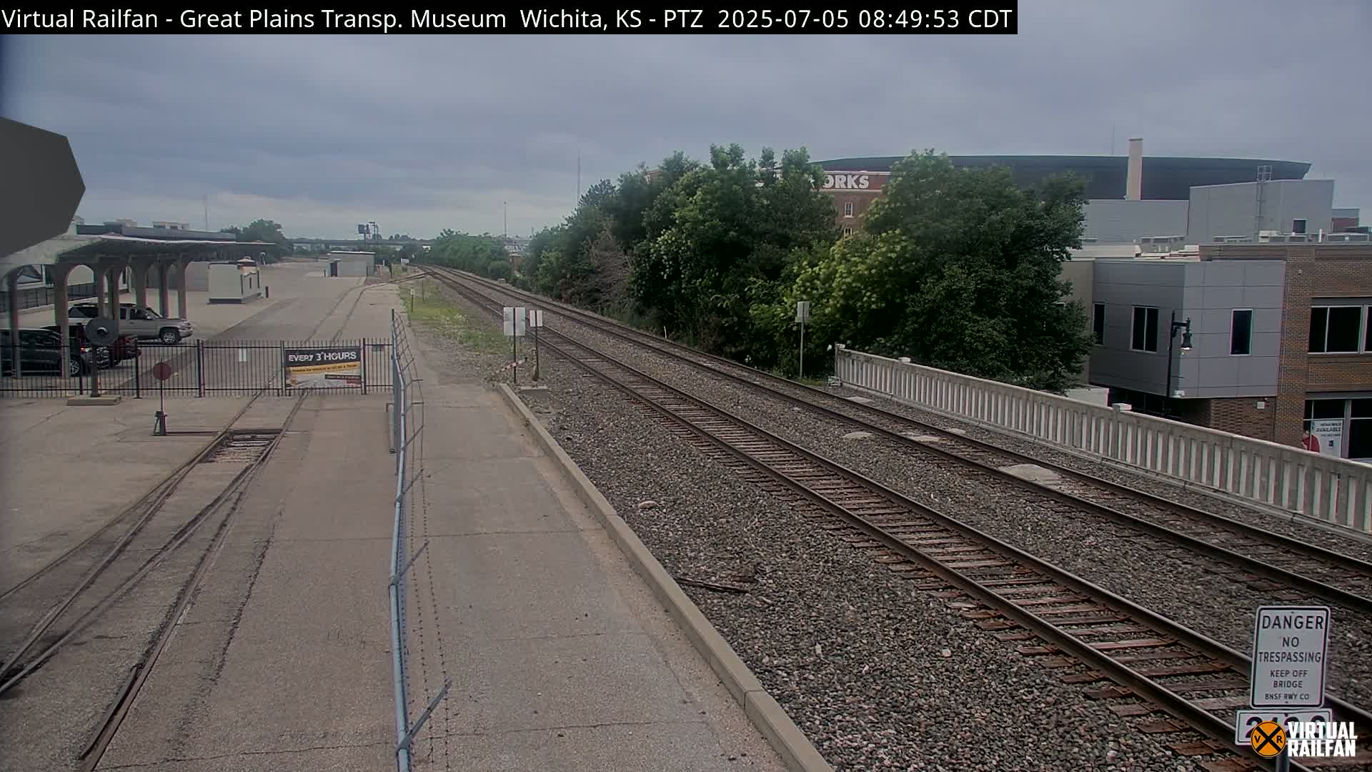 A view of railroad tracks on an overcast day, extending from a paved area with vehicles parked nearby to a bridge over a road, with buildings and trees in the background.