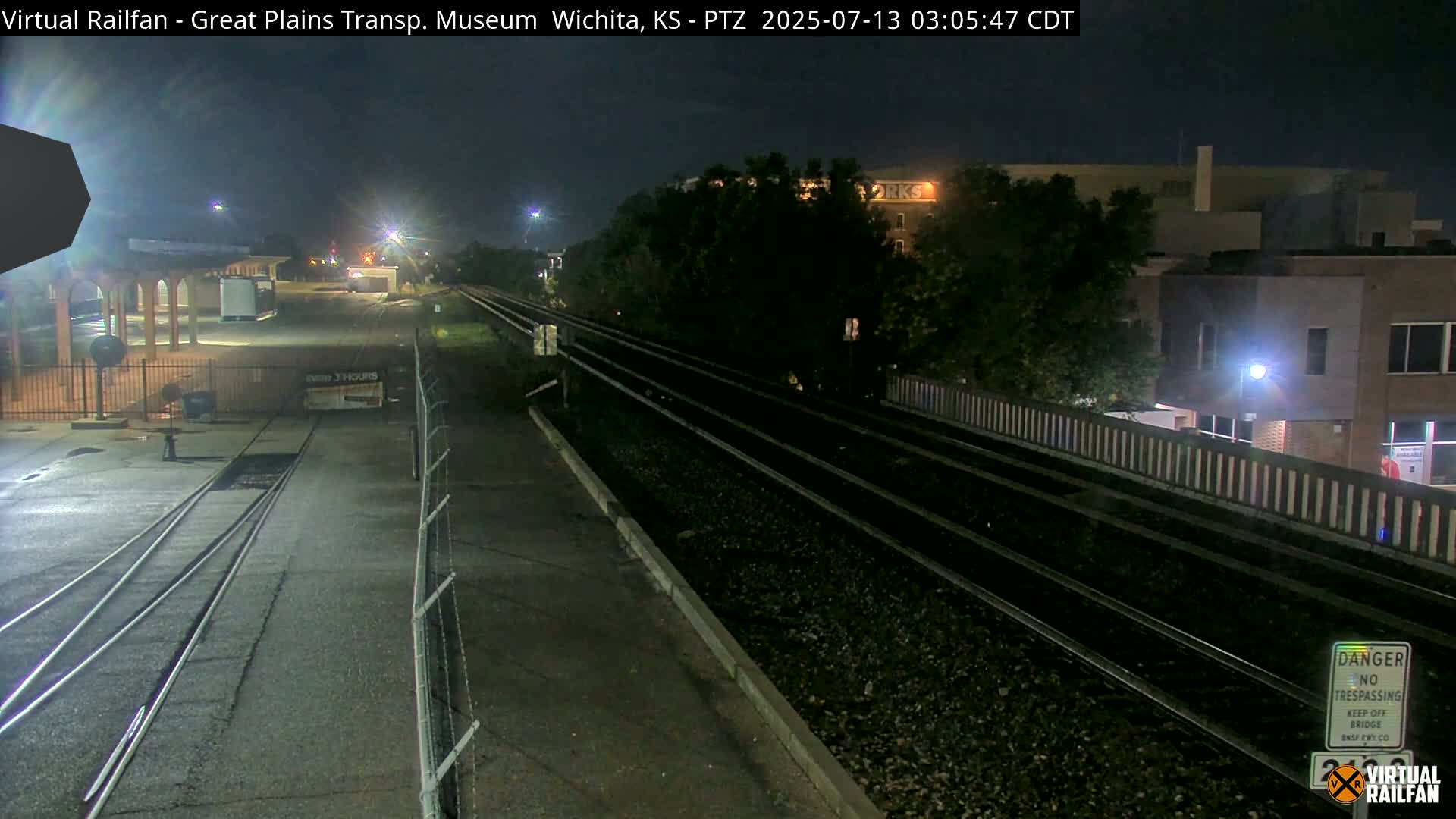 A nighttime view of train tracks beside a paved road and fenced-in area, with buildings and trees visible in the background under a dark sky.