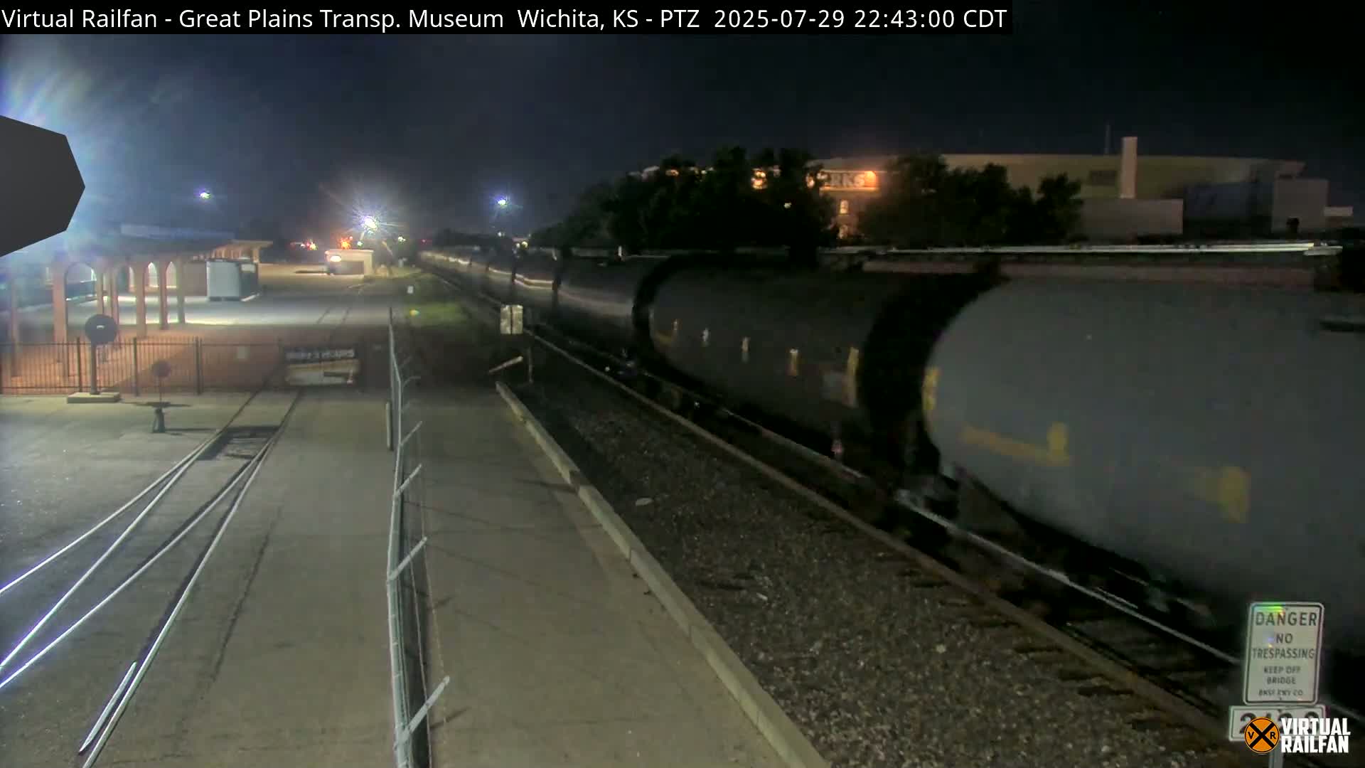 At night, railroad tracks extend into the distance past a building and some trees under a clear, dark sky.