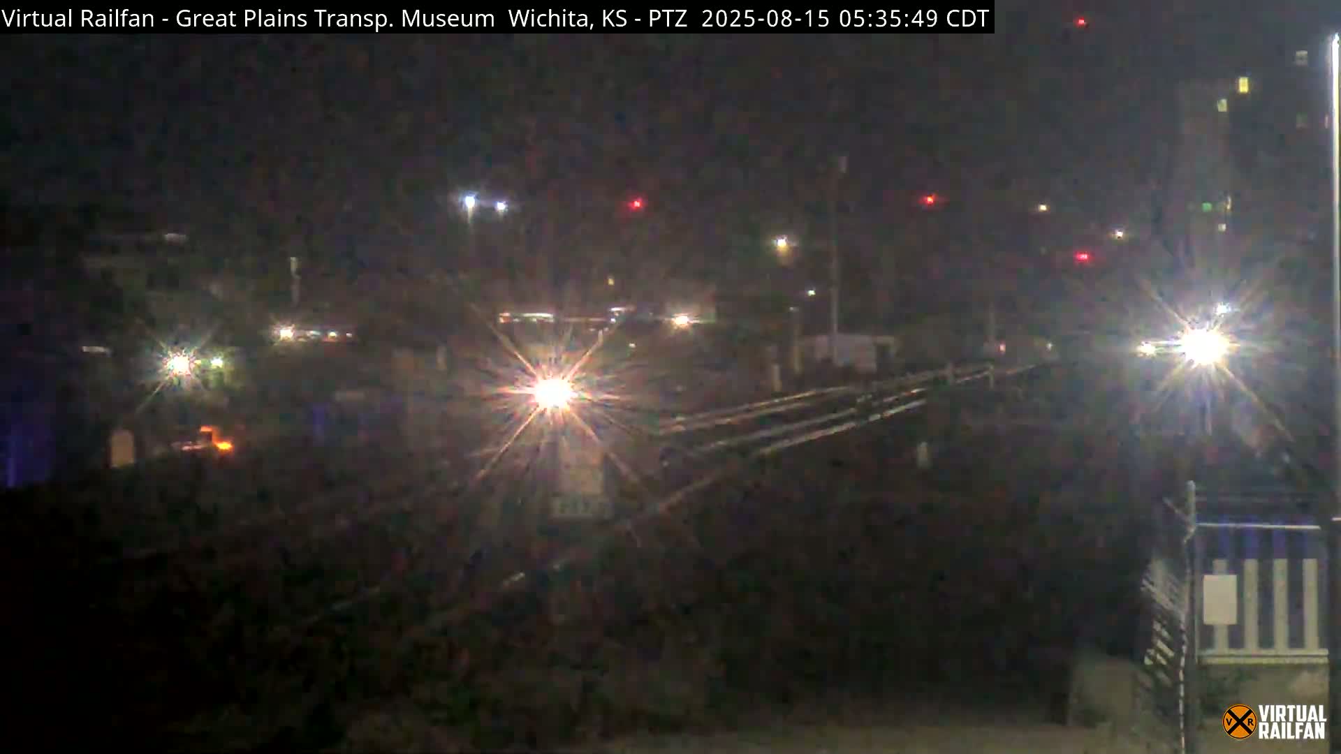 At night, railroad tracks extend into the distance past a building and some trees under a clear, dark sky.