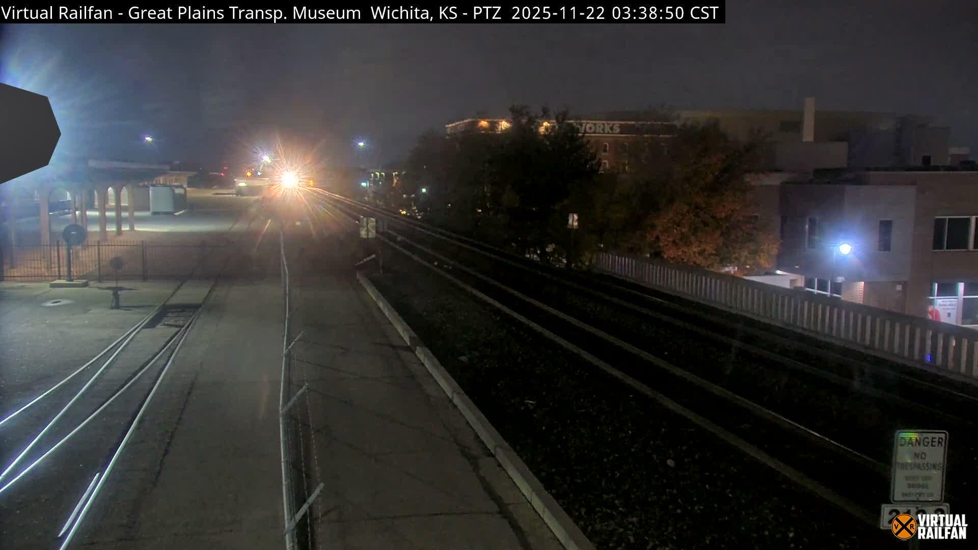 At night, a distant train with a brilliant headlight approaches on multiple railway tracks, bordered by trees and industrial-looking buildings beneath a clear sky.