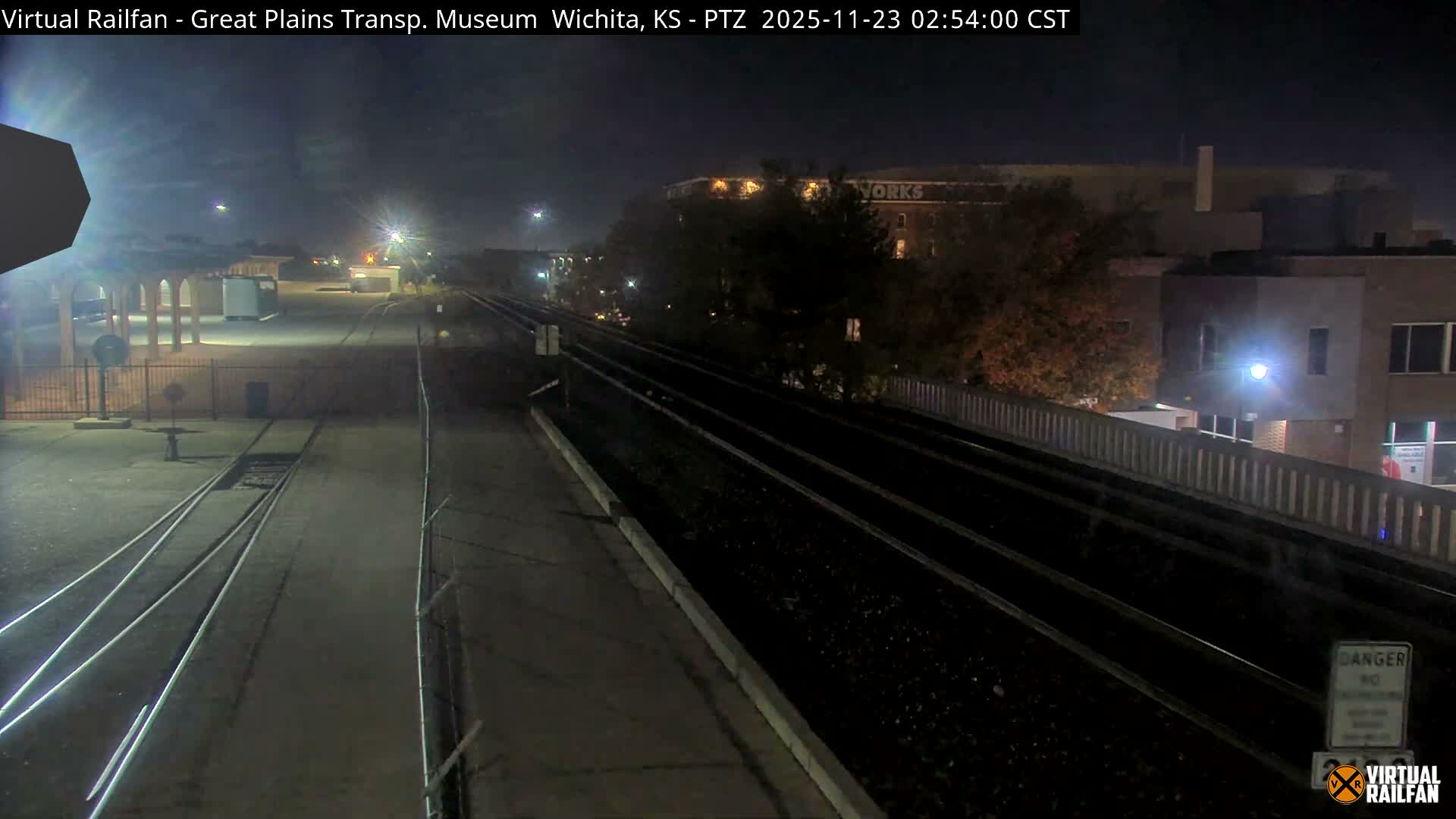 A nighttime view of a railway yard features multiple parallel tracks and converging lines alongside concrete platforms, with various industrial buildings and trees illuminated by artificial lights under a clear sky.