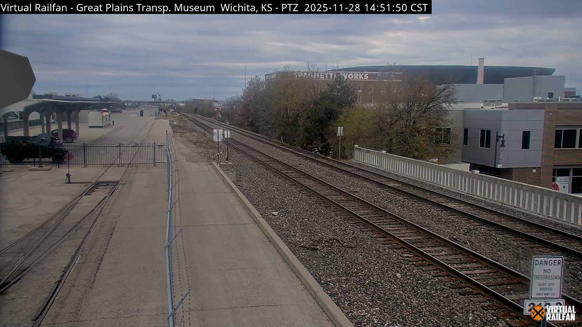 Multiple railroad tracks stretch into the distance under an overcast sky, flanked by a rail yard with a pavilion and parked vehicles on the left, and various buildings with autumn trees on the right.