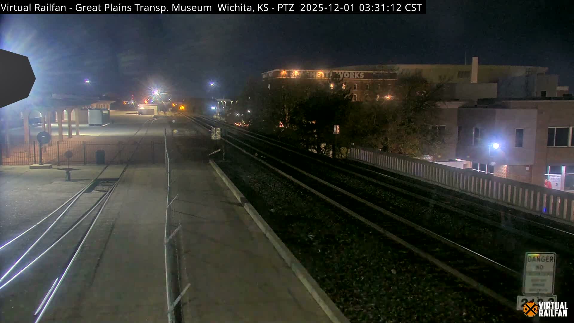 A clear night view shows multiple train tracks stretching through an urban rail area, bordered by a covered platform with columns on the left and various buildings with lit windows in the background.