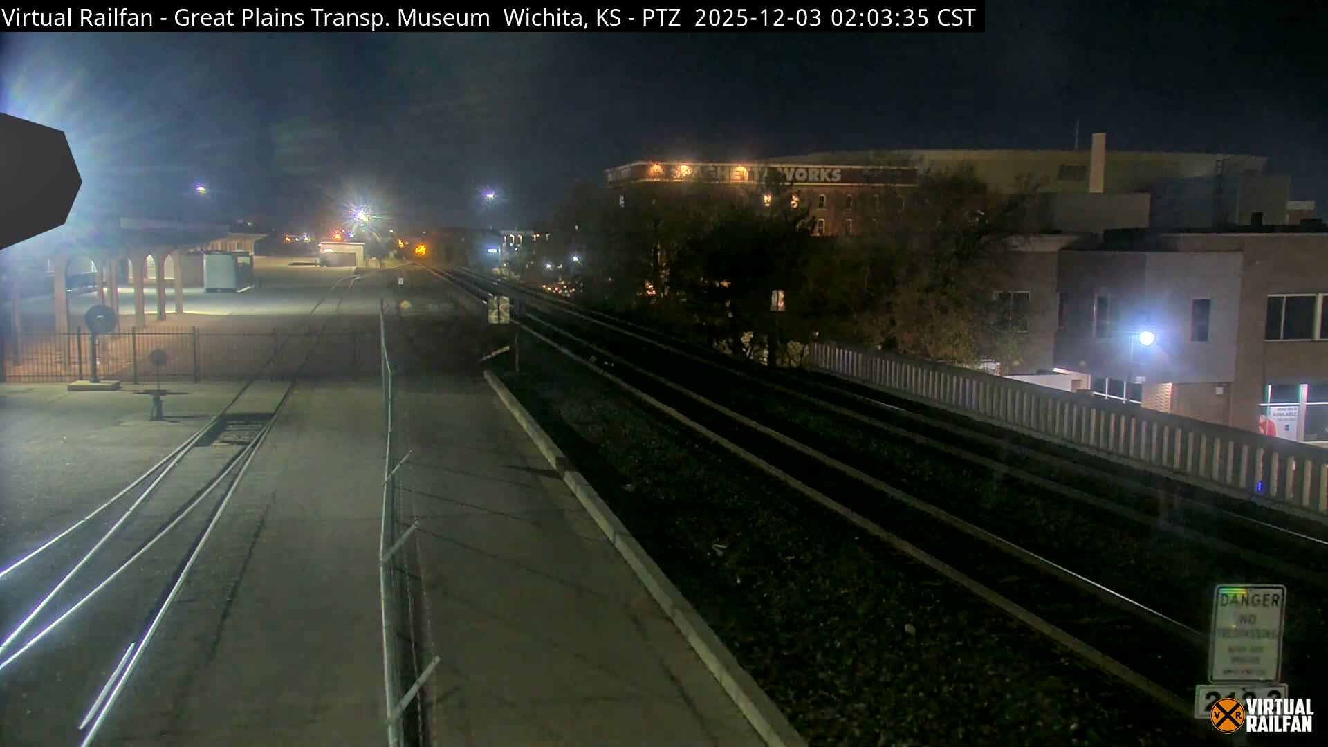 A nighttime view reveals a complex of railway tracks and platforms illuminated by bright lights, with various buildings in the background under a clear sky.