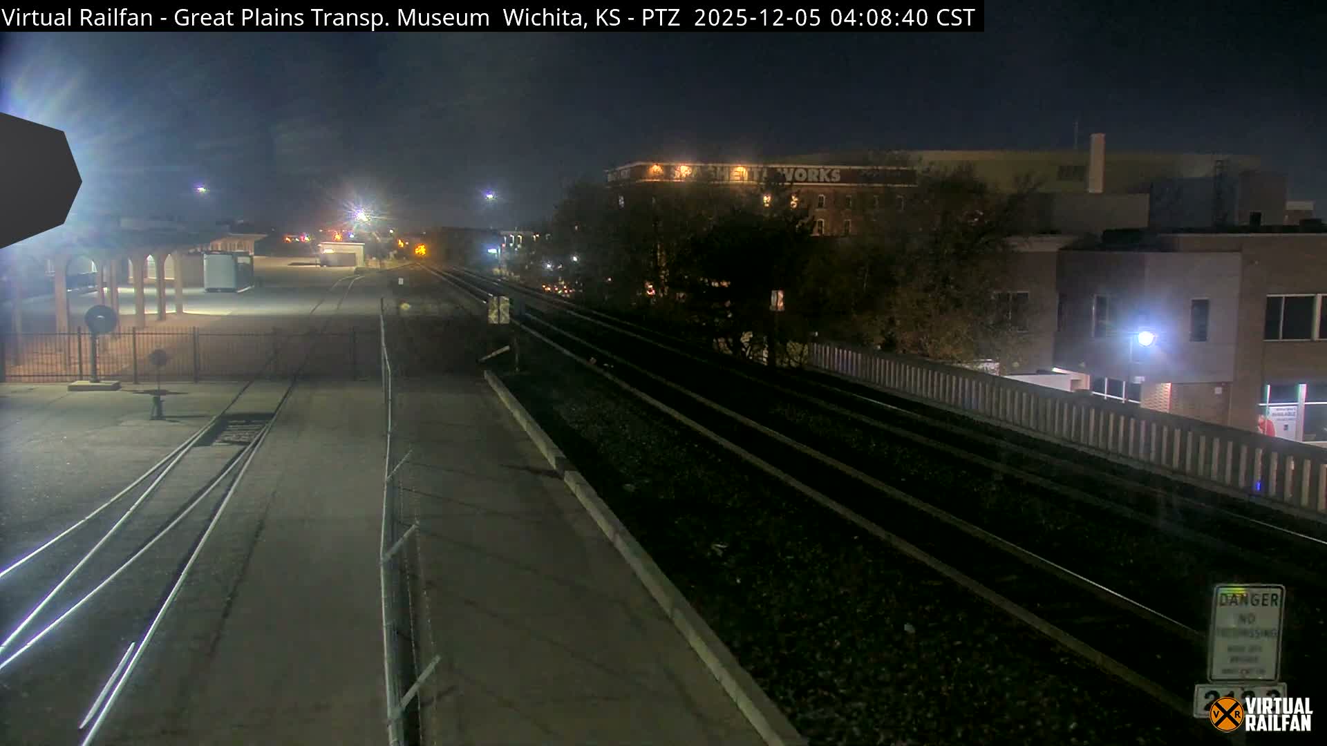 A clear, dark early morning view captures a railway area with multiple tracks, illuminated buildings, and bare trees under artificial light.
