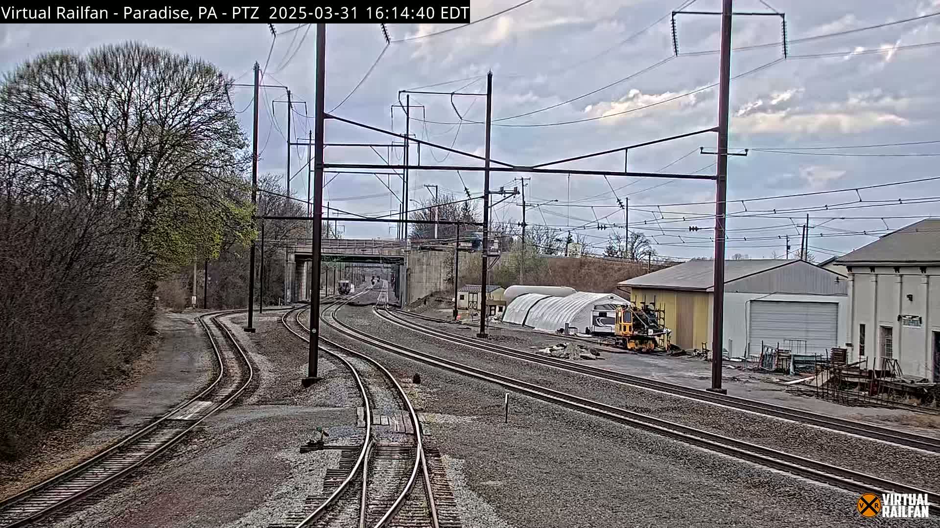 Several sets of train tracks curve through a yard with industrial buildings under a cloudy sky.