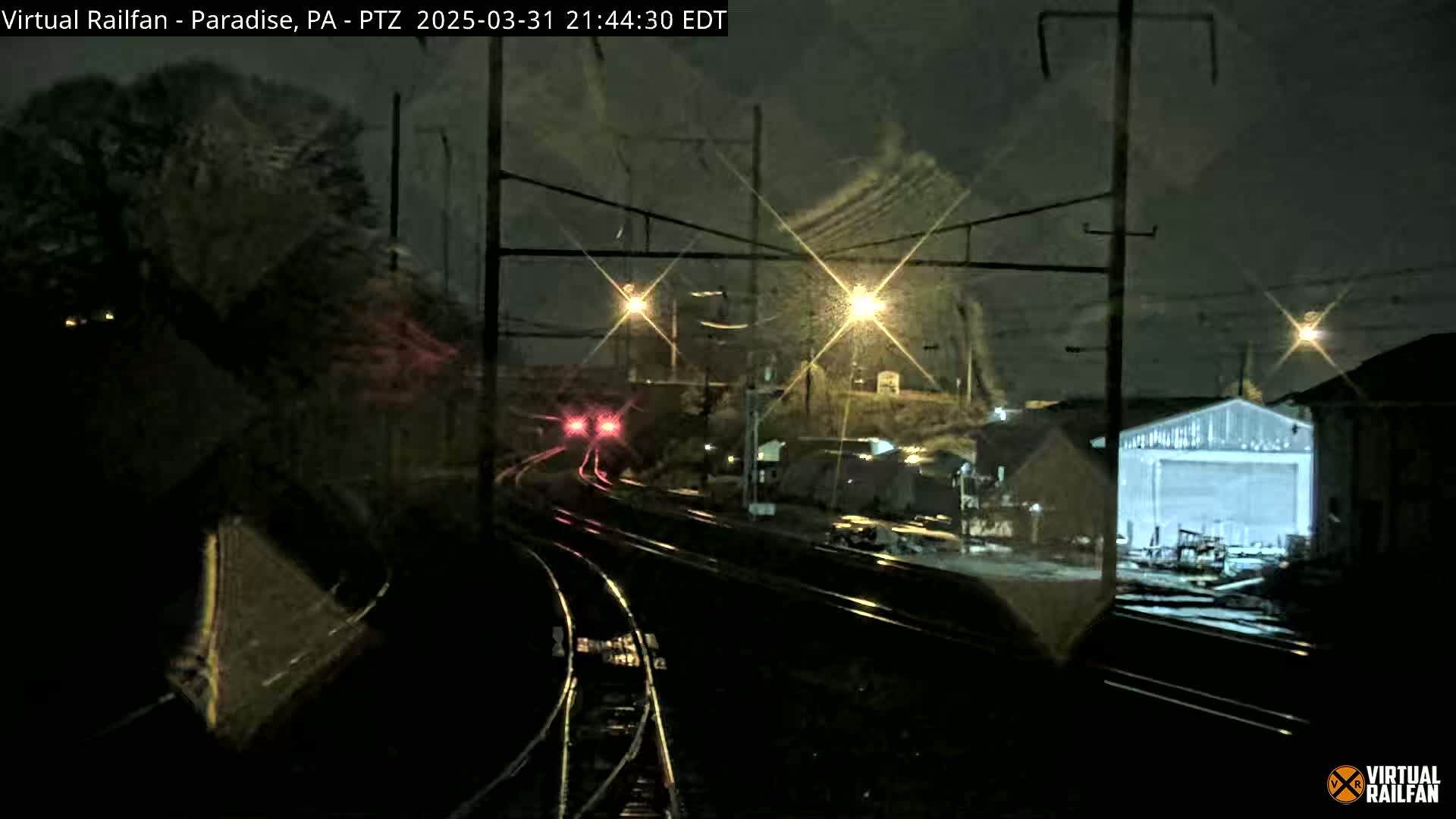 A nighttime view of railroad tracks, with a train's red tail lights visible in the distance, under snowy conditions.
