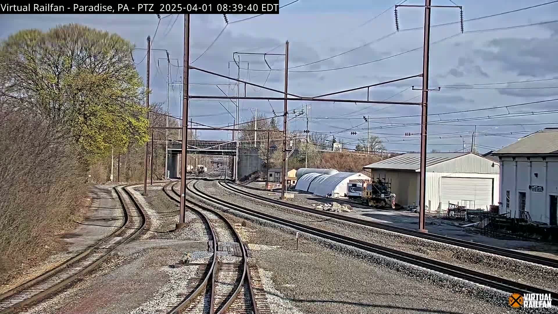 Multiple sets of train tracks curve through a landscape under a partly cloudy sky, passing near some buildings and a large tree showing early spring leaves.