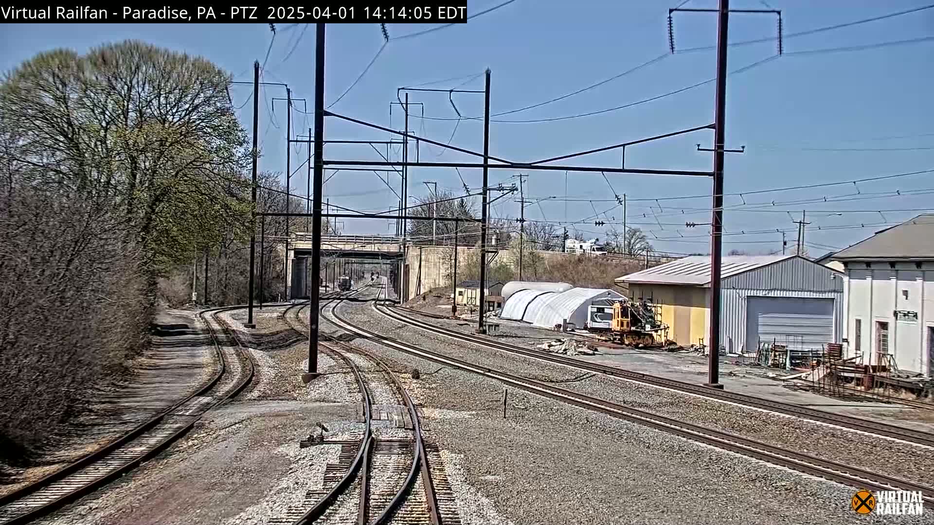 Several sets of train tracks curve through a landscape of trees and industrial buildings under a clear blue sky.