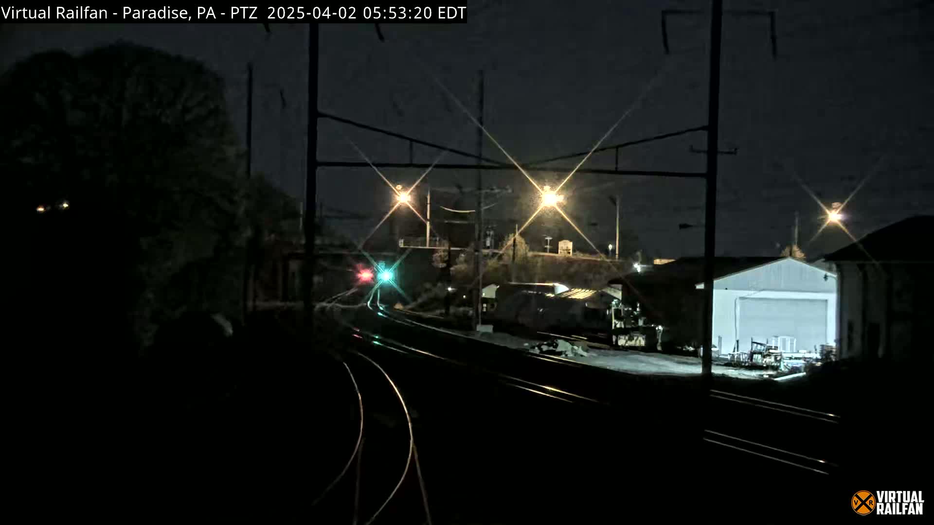 At night, a dark train sits alongside train tracks near a building, illuminated by several bright lights.