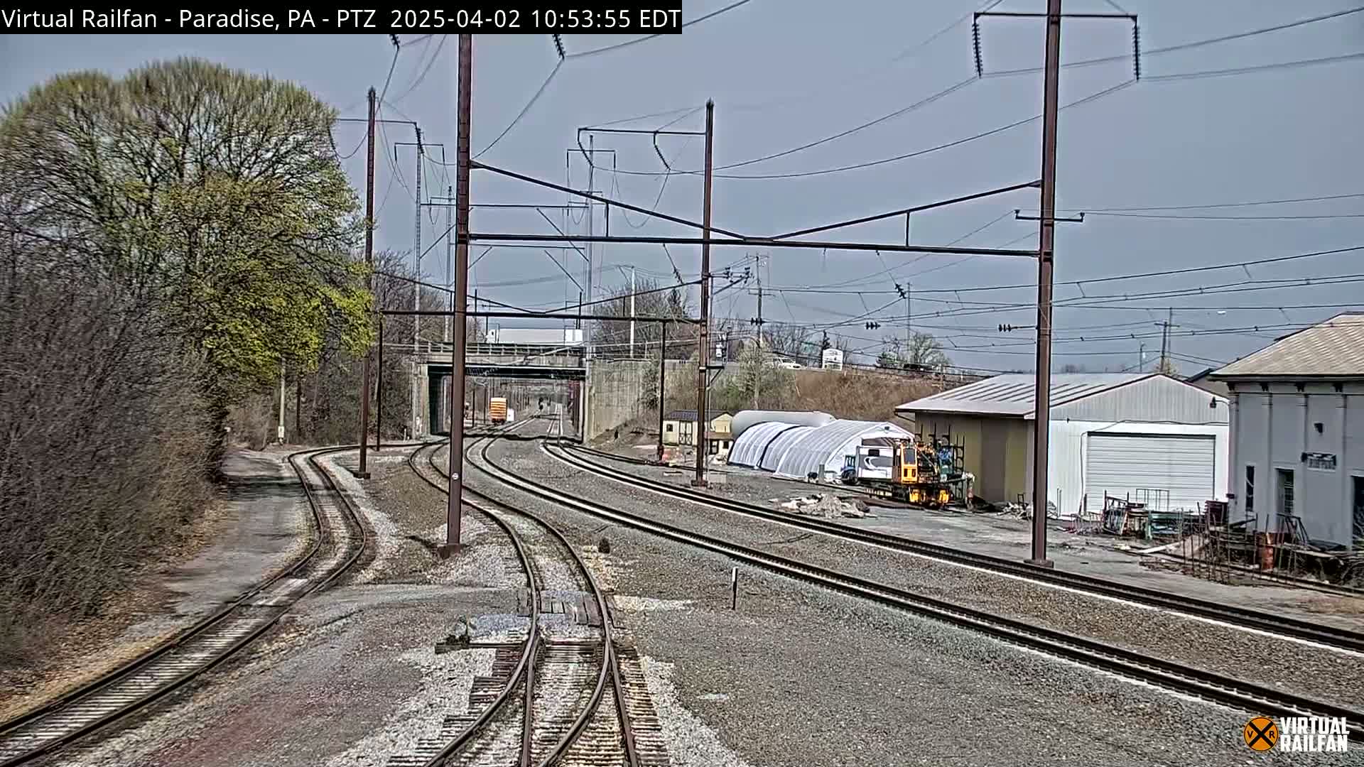 A view of several train tracks branching off from each other under a partly cloudy sky, with trees and industrial buildings in the background.