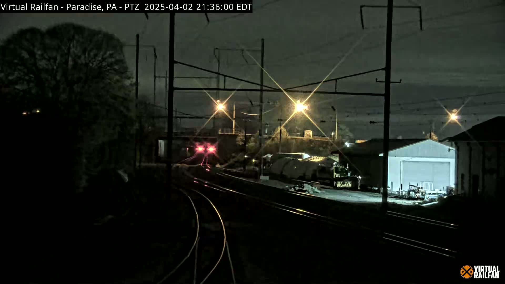 A nighttime view of railroad tracks with a train in the distance, under a dark sky with lights illuminating the area.