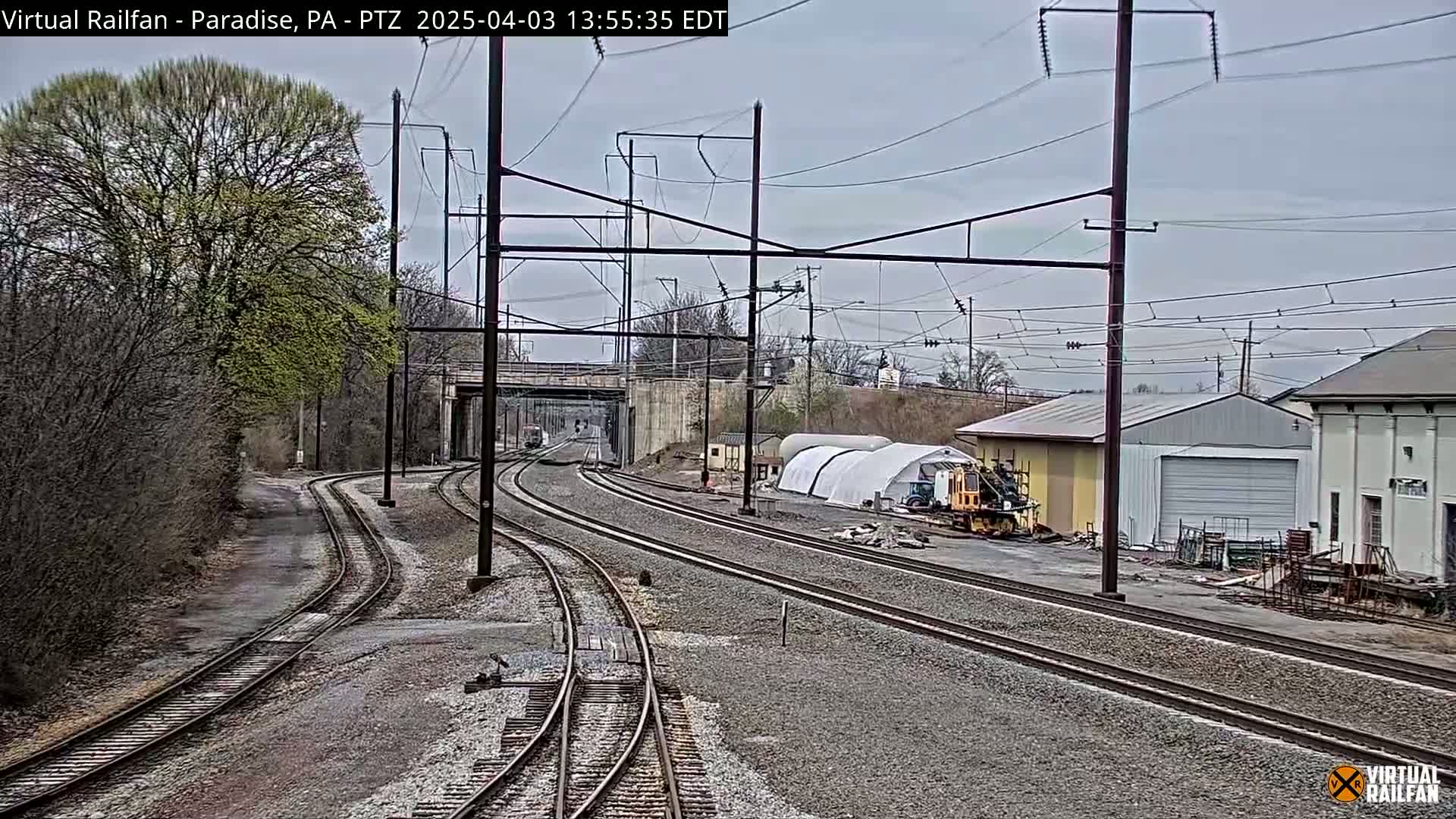 Multiple sets of train tracks converge near some buildings and a temporary structure under an overcast sky.