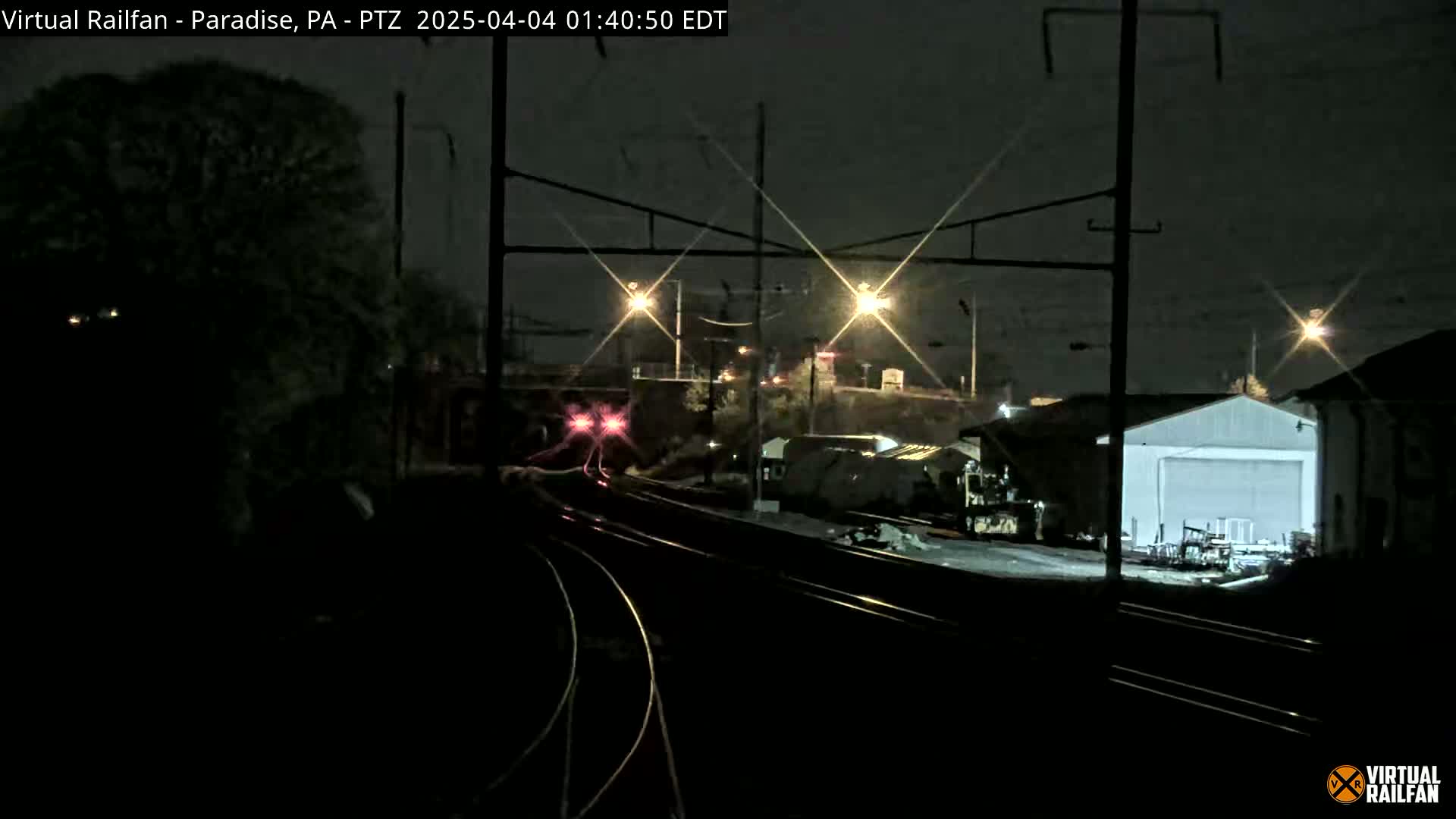 A nighttime view of railroad tracks with a train approaching, illuminated by lights and under a dark sky.