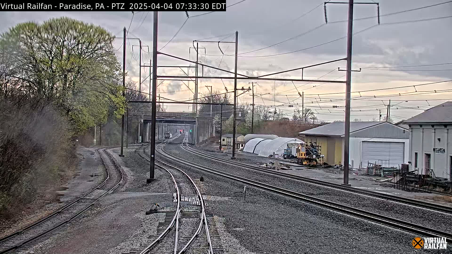 Multiple sets of train tracks extend into the distance under an overcast sky, passing alongside buildings and vegetation.