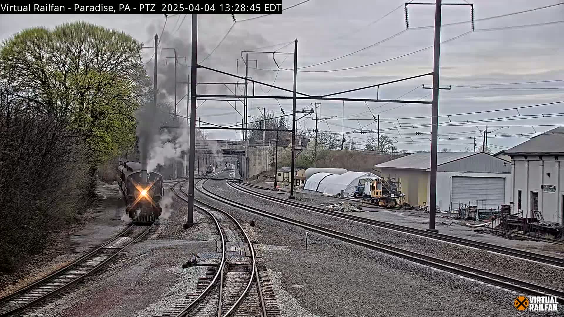 A steam locomotive is traveling on a railway track under an overcast sky, passing industrial buildings and construction equipment.