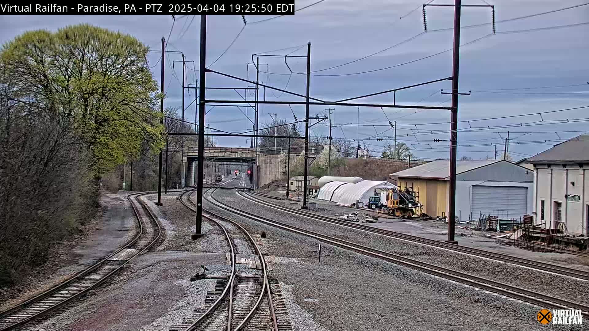 Converging train tracks under a cloudy sky pass near a road and industrial buildings, with budding trees lining the sides.