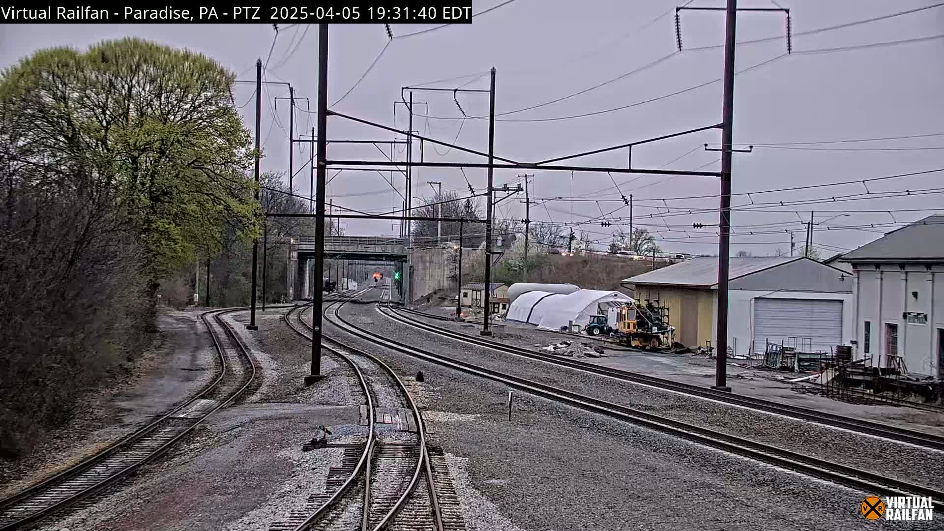 A view of several train tracks curving away from the camera under an overcast sky, with trees, buildings, and construction equipment visible in the background.