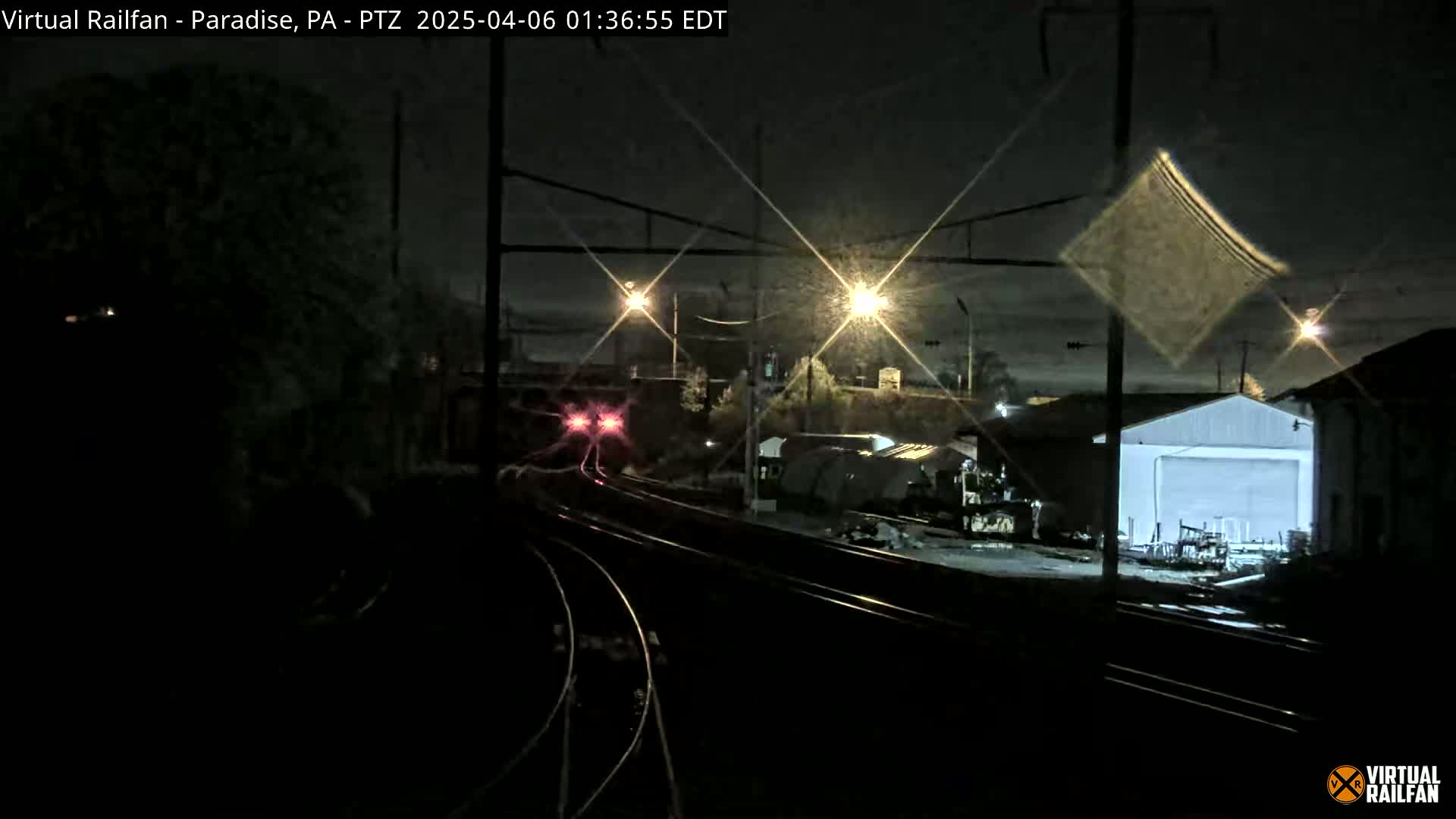 At night, a train with red lights approaches a railway junction near some buildings under a dark, possibly rainy sky.
