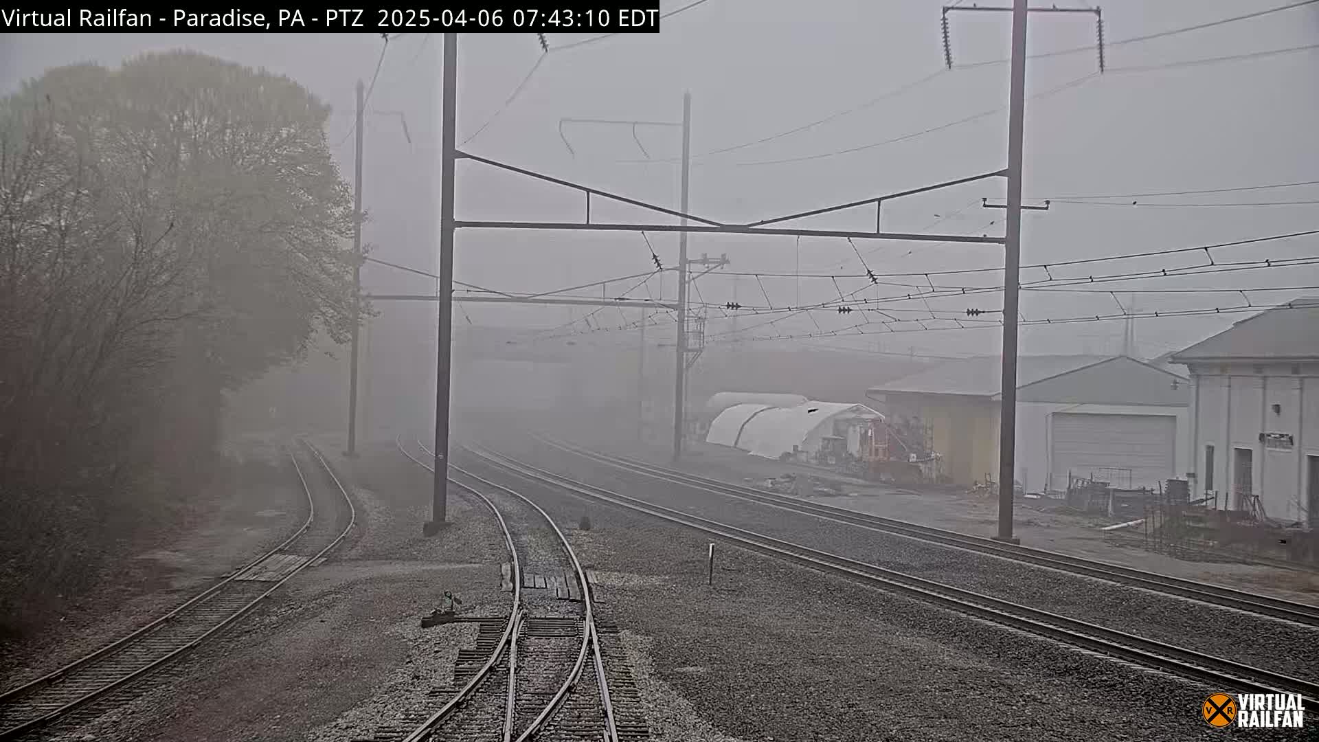 Several sets of train tracks diverge in a foggy landscape, passing industrial buildings and trees.
