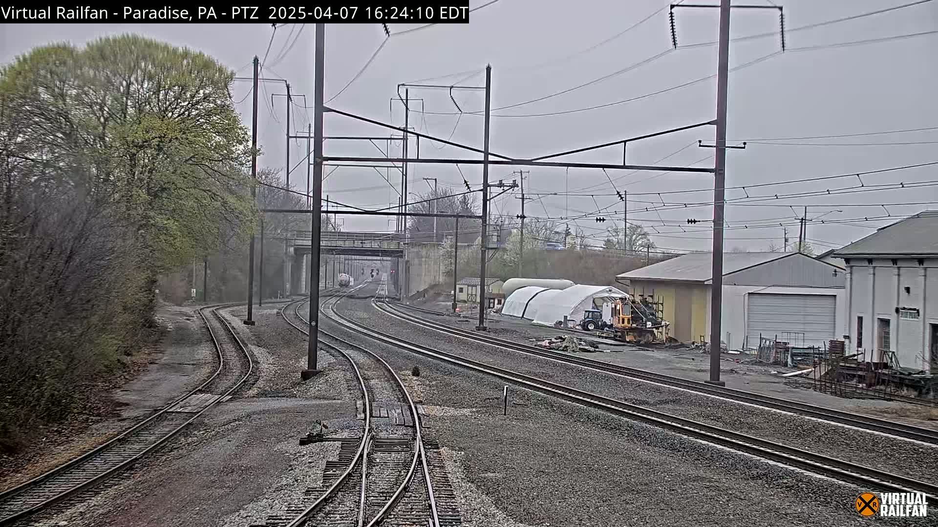 A view of railroad tracks on an overcast day, with trees, buildings, and construction equipment visible in the background.