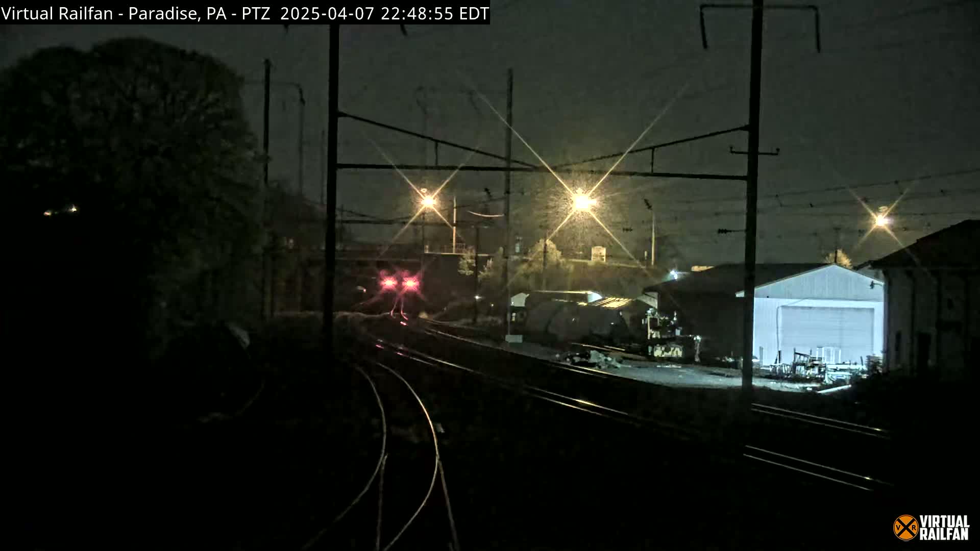 A nighttime view of train tracks with a train approaching, lit by streetlights in snowy conditions, alongside industrial buildings.