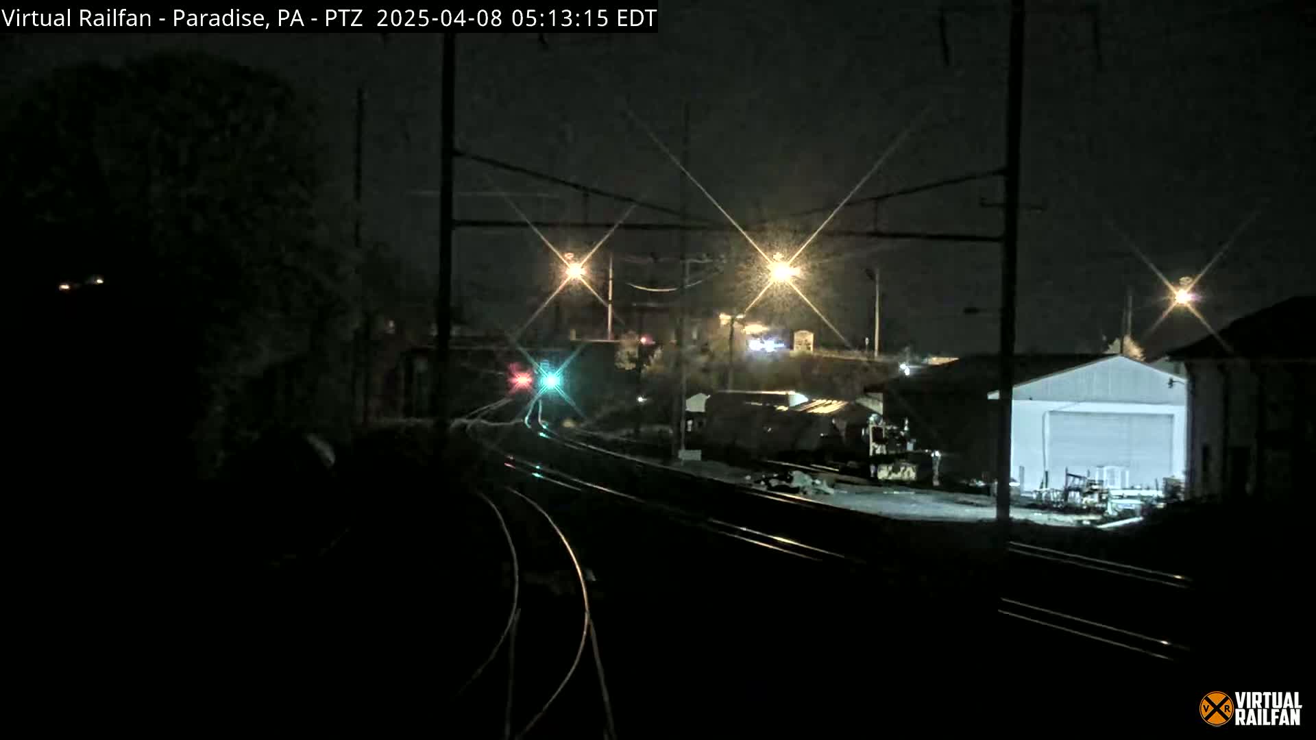 At night, a train approaches a rail yard with a large white building under bright lights.