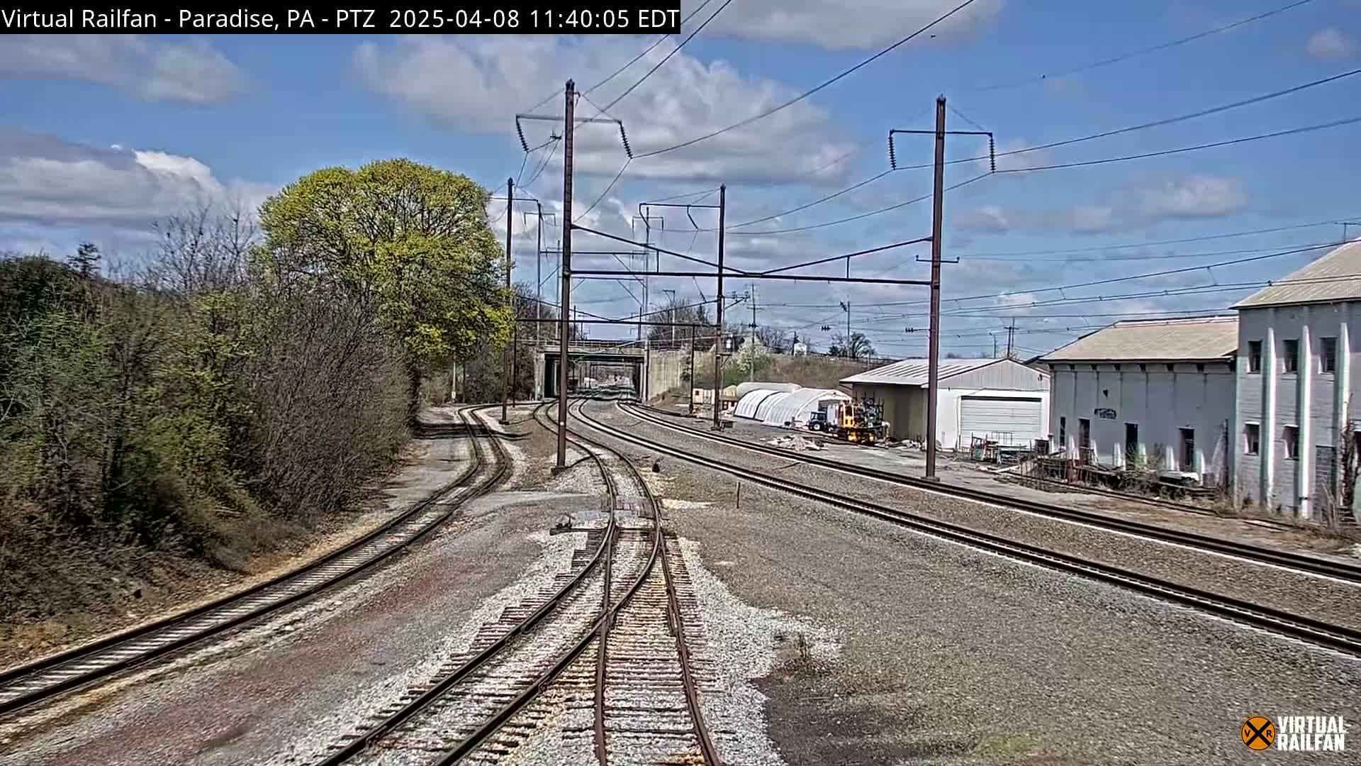 A sunny day shows a railroad yard with several train tracks converging, buildings, and trees with new spring leaves.