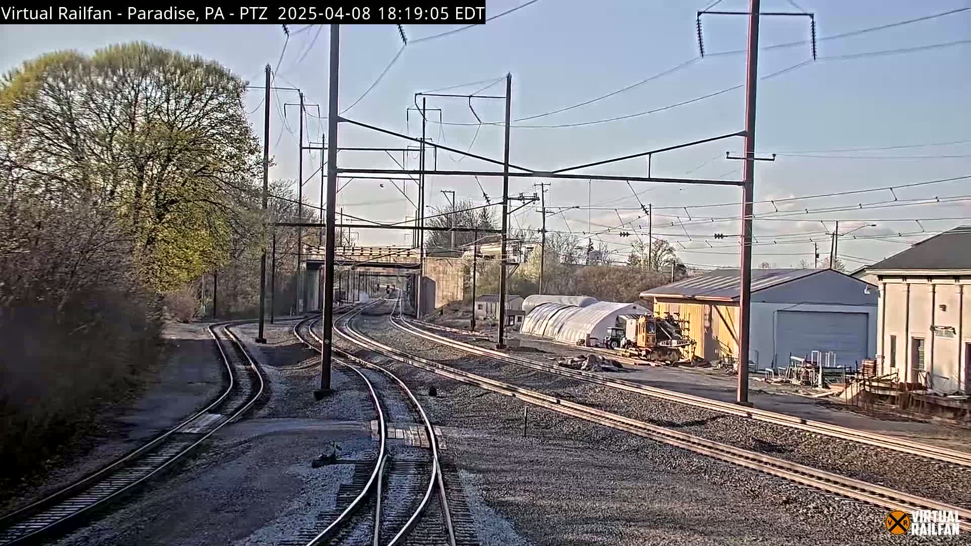 A sunny day view of several railway tracks that converge and diverge near some buildings and trees.