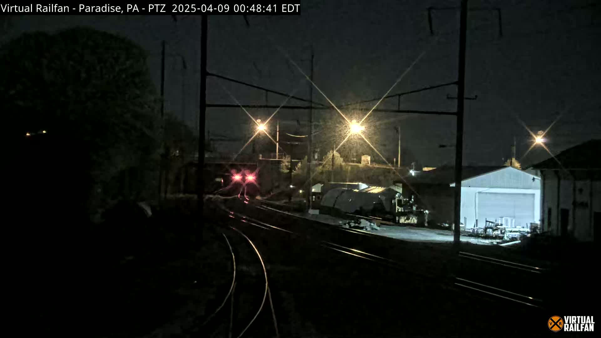 At night, under a slightly hazy sky, a nighttime view shows train tracks with a train in the distance and several buildings and lights.