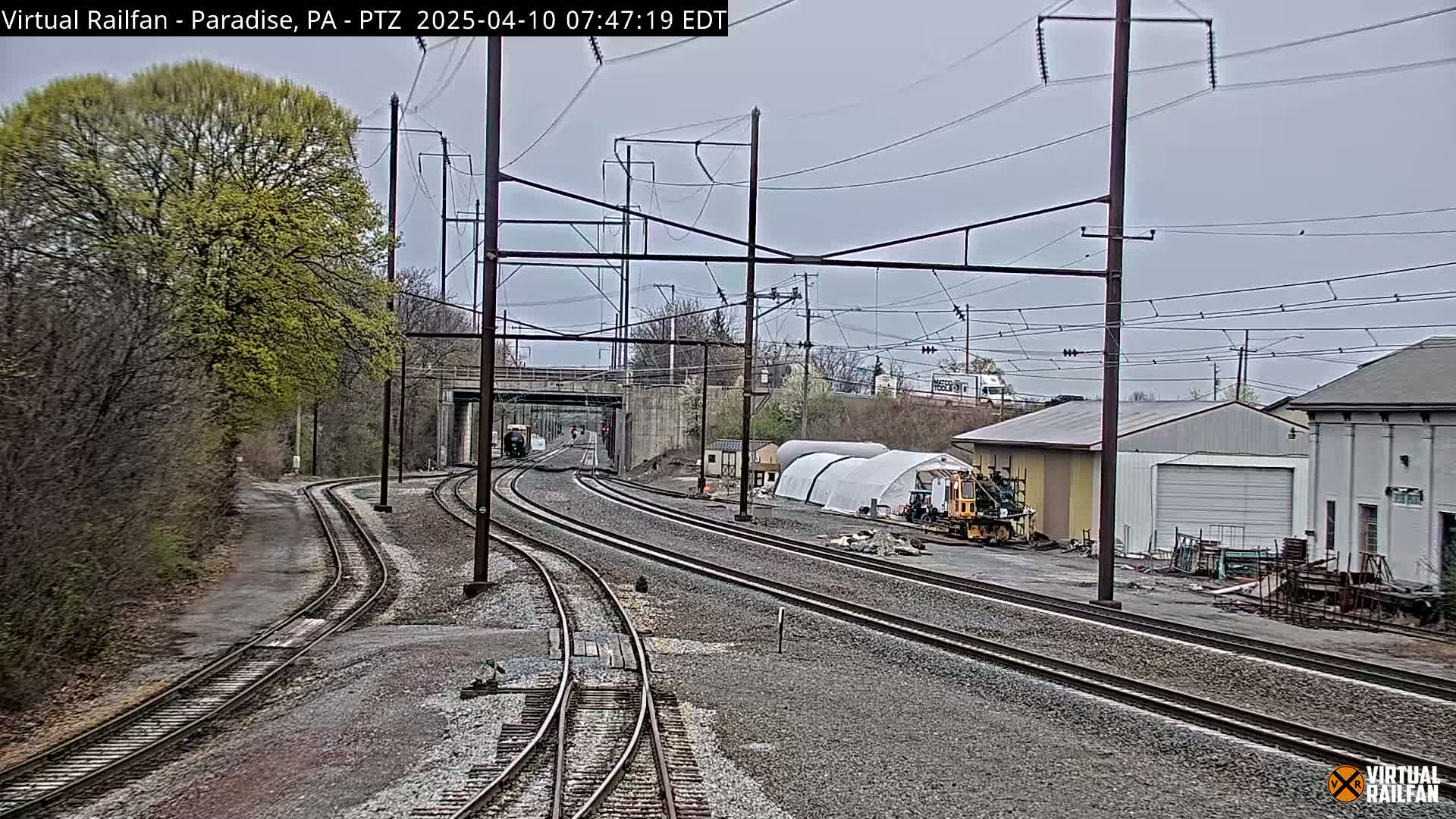 Multiple train tracks curve through a landscape of trees budding with new leaves under an overcast sky, passing near industrial buildings and a temporary structure.